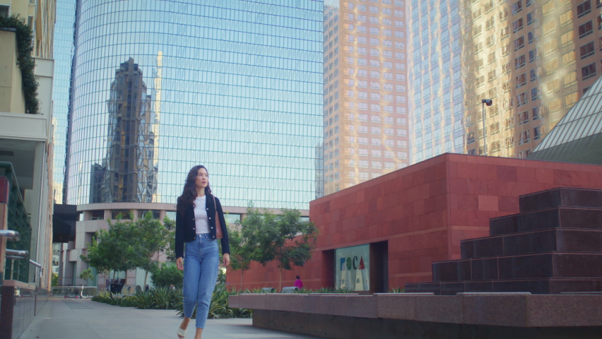 Confident businesswoman going to office on sunny day. Attractive brunette woman walking megapolis street in stylish clothes. Pretty asian lady touch hairstyle rushing to meeting in business centre.