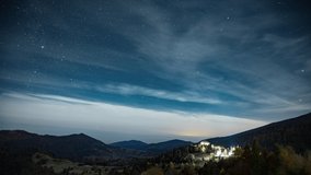 Night starry sky with white clouds floating above small town illuminated by street lights in highland near mountains 8K time-lapse