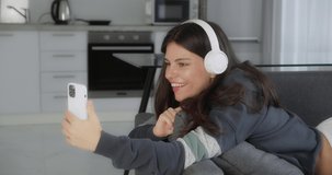 portrait of teen girl communicating by video chat in smartphone, lady with headphone is sitting on couch in modern apartment