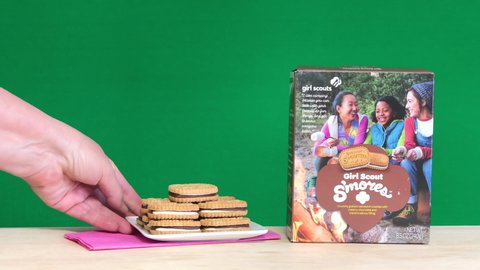 Alameda, CA - Feb 10, 2022: 4K HD video one box of S'mores Girl Scout cookies on a light wood table, hand reaches in and places pink napkin then plate with fresh cookies. Green background
