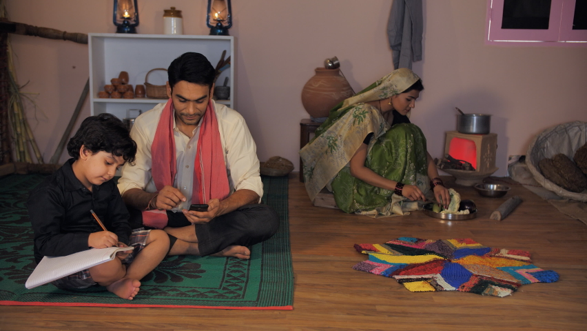 Small nuclear family in an Indian village - Indian man speaking on phone. Young kid studying in a village home. A village woman in a traditional Sari cutting vegetables while working in the kitchen.