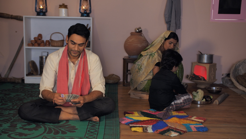 Small nuclear village family in Indian - Indian village home. Young parents and their kid. A happy farmer in casual clothes counting his hard-earned money while talking on a mobile - savings