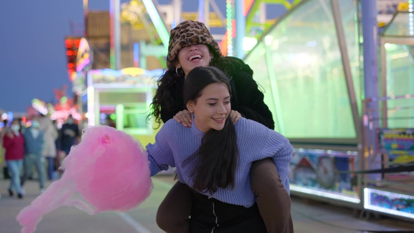 One girl gives another a piggyback ride holding pink candy floss. Best friends teenagers having fun at the funfair at night - slow-motion Royalty-Free Stock Footage #1086808232