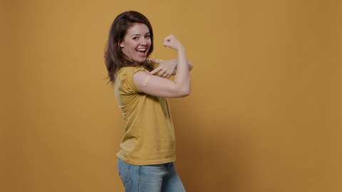 Portrait of smiling woman lifting sleeve and showing band aid after covid or flu vaccine flexing arm biceps in studio. Proud person revealing bandage covering immunization spot for medical awareness.