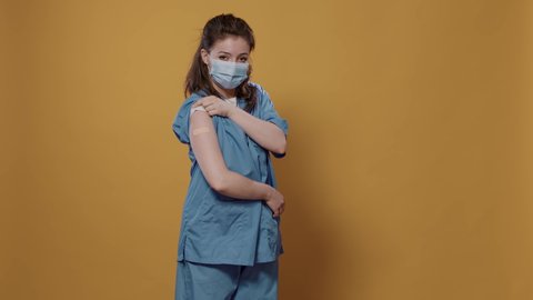 Portrait of medic wearing surgical mask lifting sleeve and showing band aid after covid or flu vaccine in studio. Medical doctor in uniform revealing bandage covering immunization spot.