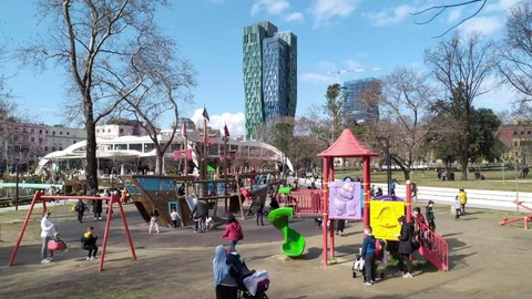 City park with playground in Tirana Albania, people enjoying sunny winter day. Kids playing and having fun on slide, swing and playhouse, a tower in the background, under blue sky. Feb 06.2022.