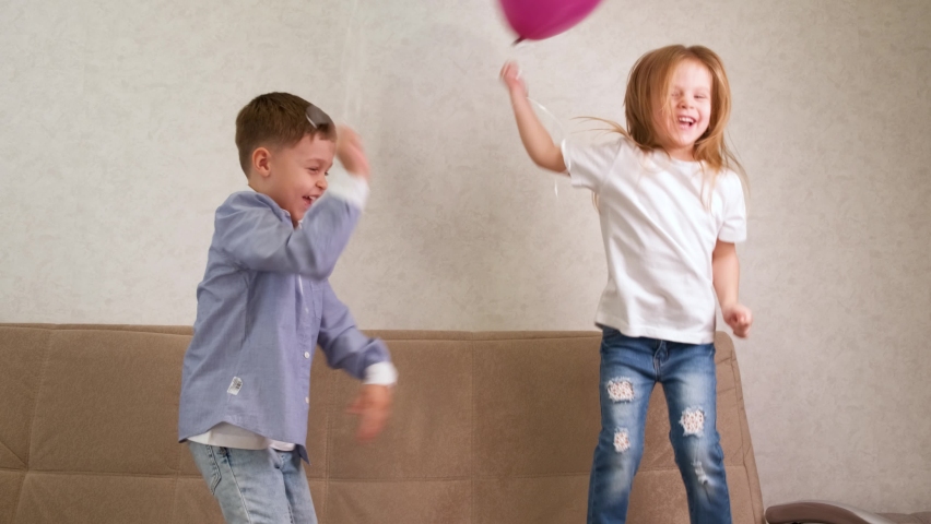 A little girl and a boy are jumping and having fun on the couch with blue and purple balloons. Happy carefree childhood. Children rejoice