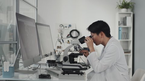Tracking shot of Asian female lab technician using microscope while African American electronics engineer working with multimeter and computer at desk