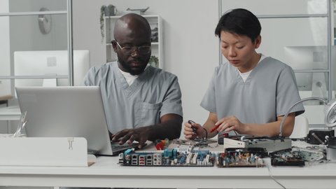 Asian female engineer using multimeter and talking with African American colleague as he working on laptop at desk in laboratory