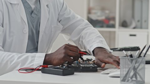 Close up tracking shot of hands of African American male engineer using multimeter and typing on computer while working with electronic components