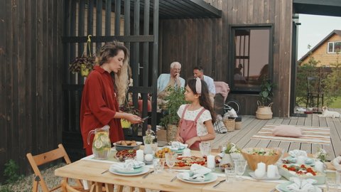Medium long of Caucasian woman and girl serving table outdoors wooden summer house, talking, two men sitting in armchairs on background on sunny summer day