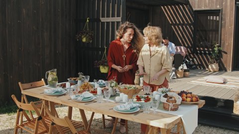 Wide shot of two younger and older Caucasian women serving table outdoors wooden summer house, putting bowls with food on it, two men sitting in armchairs on background on sunny summer day