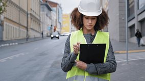 African american female engineer in safety vest helmet hardhat engineer standing in city checking road traffic pointing with finger in distance checks data in clipboard. Professional woman logistic - Powered by Shutterstock - Get 15% off with code: PIKWIZARD15