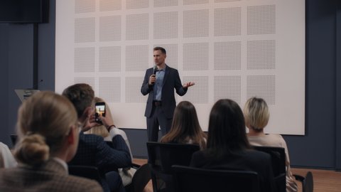 Speech of male employee at business conference for office workers. Speaker talks to audience at scene in crowded hall. Adult man explains motivation to group of people. Summit meeting at modern event Arkistovideo
