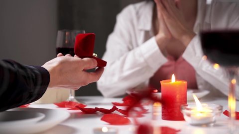 Couple dine by candlelight. Hands of lovers on table with glasses of red wine. Man proposes for Valentine's Day. Male hands hold out box with wedding ring. Marriage proposal, during romantic dinner