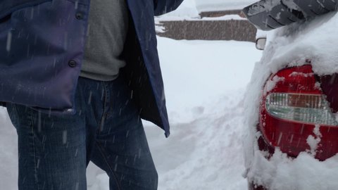A man in a jacket and jeans is shoveling snow off his car. Close-up. A red car is covered in snow during a snowstorm. Snowfall in the background