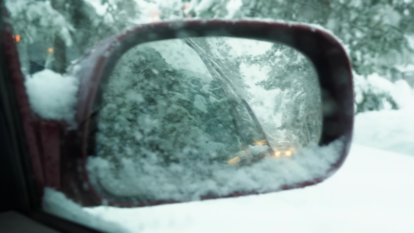 View from the car in the side rear view mirror. The car is driving through a snow-covered city. In the background are trees and snowfall