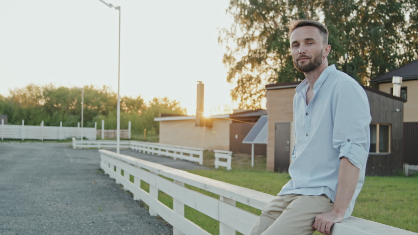 Medium long of blue-eyed Caucasian man wearing casual clothes, leaning on white fencing in neighborhood in summer evening, smiling and looking on camera