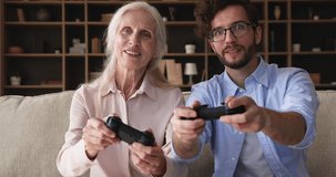 Mature grandmother and young adult grandson having fun together at home sit on sofa holding gamepads play racing video games, celebrate victory giving high five feel excited. Hobby, amusement concept