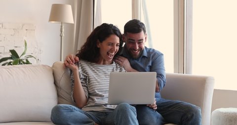 Beautiful millennial couple sit on sofa use laptop search information find last minute travel tour offer hugging feel overjoyed, online lottery win, success, achievement, auction victory, luck concept