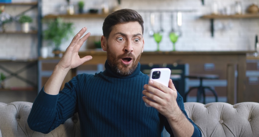 Excited and pleasantly surprised handsome man rejoices at the message on his smartphone. Young male looking at mobile phone and feel amazed happy and saying WOW on home background. Royalty-Free Stock Footage #1086827798