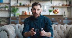 Young man with beard playing console video games with joystick gets upset because he losing in game while sitting on the couch at home