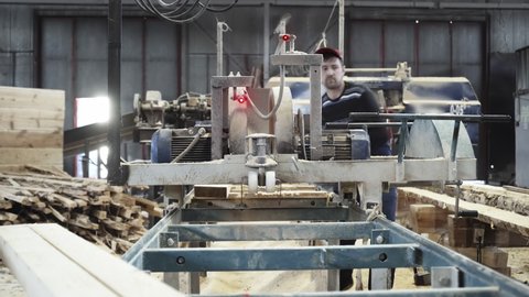 Two Male Workers Operating Industrial Saw. Industrial Saw In A Wood Production Process At Modern Timber Workshop. Industrial Saw Machine Used By Woodworkers To Cut Wooden Board Material