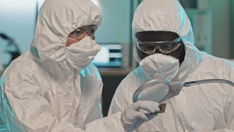 Tilt up shot of two diverse engineers in coverall suits, masks and gloves looking at computer chip under magnifying glass and having discussion while cooperating in laboratory