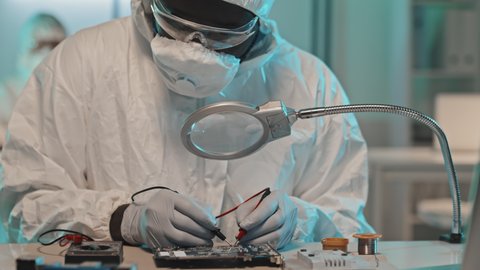 Tilt down shot of electronics engineer in coveralls, mask and gloves using multimeter while testing computer circuit board in laboratory