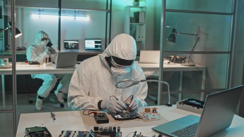 Tilt up shot of African American technician in coveralls, mask and gloves using multimeter while testing computer chip in laboratory