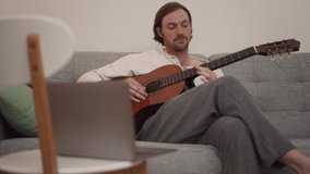 Pensive long-haired man learns to play the guitar from a video lesson at home