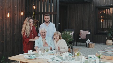 Medium long of younger Caucasian couple standing behind senior parents who sitting at table in backyard in summer evening, video calling via tablet computer