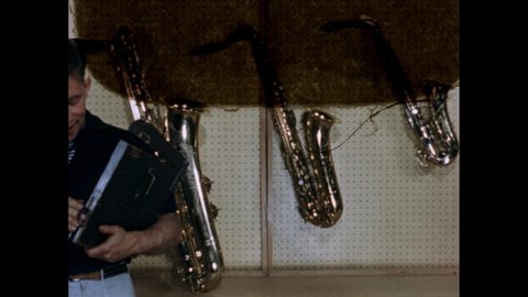 1950s: Man holding clapperboard. Saxophones hanging on peg board. Instruments on wall. Clapperboard clapping. Man sitting on stage, inserting reed into oboe, playing oboe. Musician playing instrument