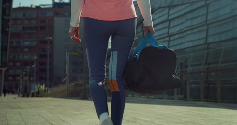 Cinematic close up shot of young millennial woman with athletic body wearing sportswear carrying bag is walking in busy city center street on way to gym for yoga or fitness workout training activity.