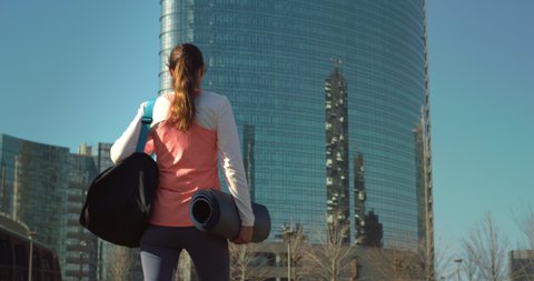 Cinematic shot of young millennial woman with athletic body wearing sportswear carrying bag and mat is walking in busy city center street on way to gym for yoga or fitness workout training activity.