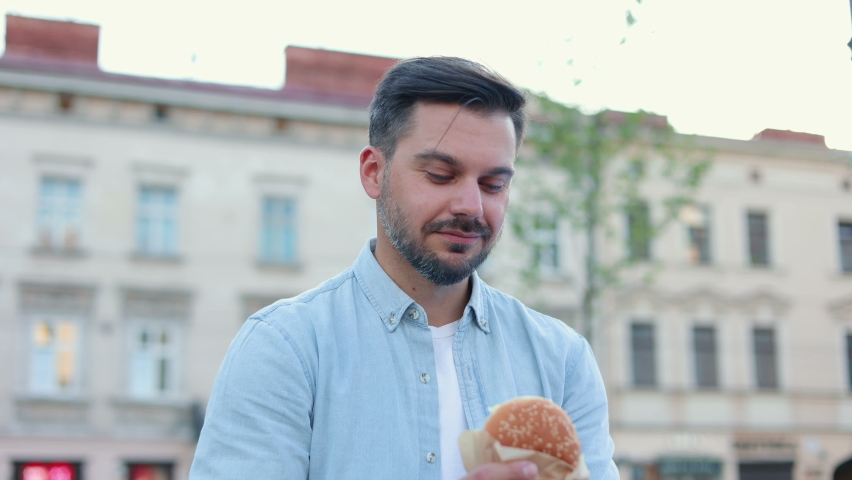 Portrait of unshaved caucasian man eating meat burger with pleasure, male casually eats junk food standing on street. European middle aged person eating american hamburger. Royalty-Free Stock Footage #1086838676