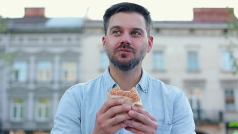Portrait of unshaved caucasian man eating meat burger with pleasure, male casually eats junk food standing on street. European middle aged person eating american hamburger.