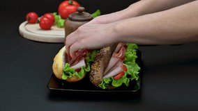 Video with woman hand preparing tasty sandwiches with ham, cheese and vegetables in black plate, close up view	