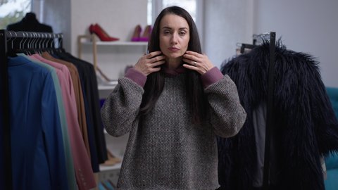 Medium shot portrait of confident Caucasian beautiful woman trying on oversized sweater with hood in fashion shop indoors. Front view thoughtful female buyer choosing new clothing in slow motion