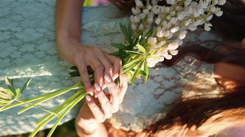 A young attractive beautiful brunette woman in a blue dress picks white lupine flowers in the middle of a flowering field and makes a bouquet of lupines. Vertical video