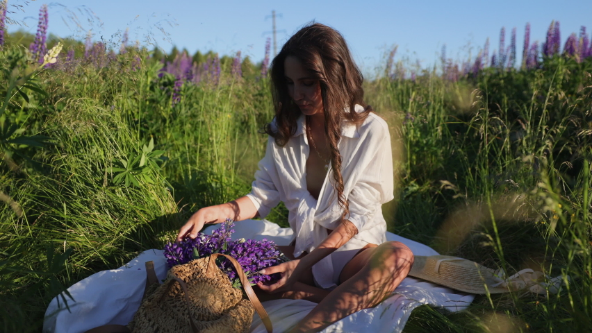 Attractive woman brunette girl sitting in a white blouse shirt on the grass in the middle of the field. Enjoying and relaxing in nature. Breathing in the scent of blooming lupines