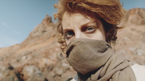 Portrait arc shot of young woman with scarf covering face looking camera while standing in post-apocalyptic desert