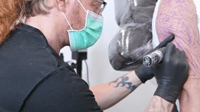 Tattooist with his machine tattooing on the arm with the design drawn and using protective mask against covid 19, concept of art and design during the new normal. High quality 4k footage