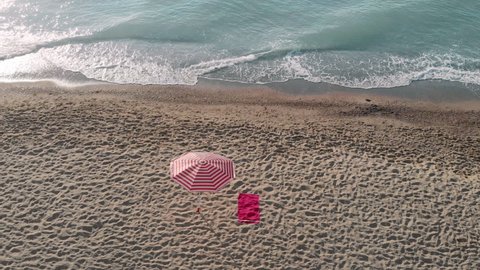 Red towel and striped sun protection umbrella on beach in summer. Sea waves wash soft sand on sunny day. Beach essentials on resort aerial view