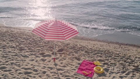 Striped beach umbrella fixed in soft sand near towel and rubber ring. Bright sun path reflecting in clear sea water. Beach essentials at resort aerial view