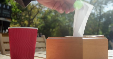 Man puts red paper cup on table in street cafe on sunny day. Hand takes clean napkin out of wooden case on blurred background closeup slow motion