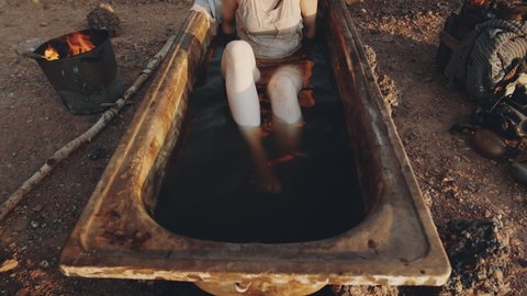 Arc shot of young woman lying in old rusty bath filled with dirty water at campsite in post apocalyptic world