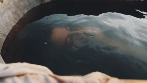 Young woman lying under dirty water in old rusty bath and then getting up at sunset in dystopian world