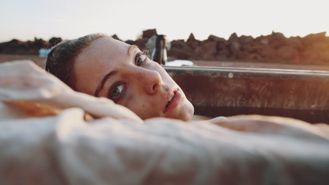 Portrait of young woman lying in rusty bathtub filled with dirty water and looking at camera at sunset in post apocalyptic world