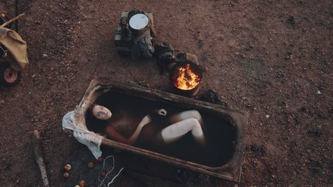 Top sown shot of young woman with pale skin lying in dirty water in old rusty bath at post apocalyptic campsite